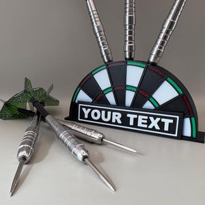 Puede incluir: Un soporte con forma de diana con tres dardos y el texto "YOUR TEXT" en blanco sobre un rectángulo negro. Varios dardos plateados están esparcidos frente al soporte, y un dardo verde y geométrico está a la izquierda. La diana tiene secciones negras, blancas y rojas.