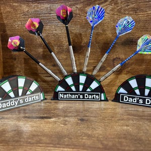 May include: Dart set with colorful flights and silver barrels, displayed in a dartboard-shaped holder. The holders are labeled "Daddy's darts", "Nathan's Darts", and "Dad's Darts". The darts are arranged on a wooden surface.