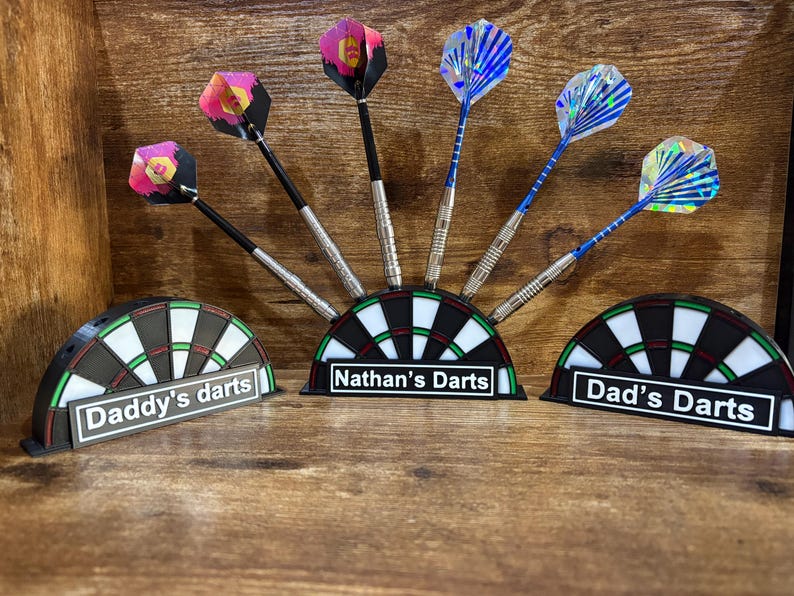 May include: Dart set with colorful flights and silver barrels, displayed in a dartboard-shaped holder. The holders are labeled "Daddy's darts", "Nathan's Darts", and "Dad's Darts". The darts are arranged on a wooden surface.