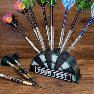 May include: A black and green dartboard-shaped dart holder with darts. The holder has a white and green target design and a rectangular space with the text "YOUR TEXT". Several darts with colorful flights are in the holder and lying on the wooden surface.