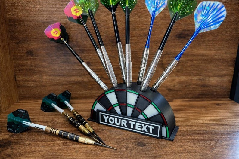 May include: A black and green dartboard-shaped dart holder with darts. The holder has a white and green target design and a rectangular space with the text "YOUR TEXT". Several darts with colorful flights are in the holder and lying on the wooden surface.