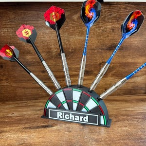 May include: A set of darts with various colorful flights, displayed in a dartboard-shaped holder. The holder is black, white, and green, with the name "Richard" on a white label. The darts have silver barrels and black or blue shafts.