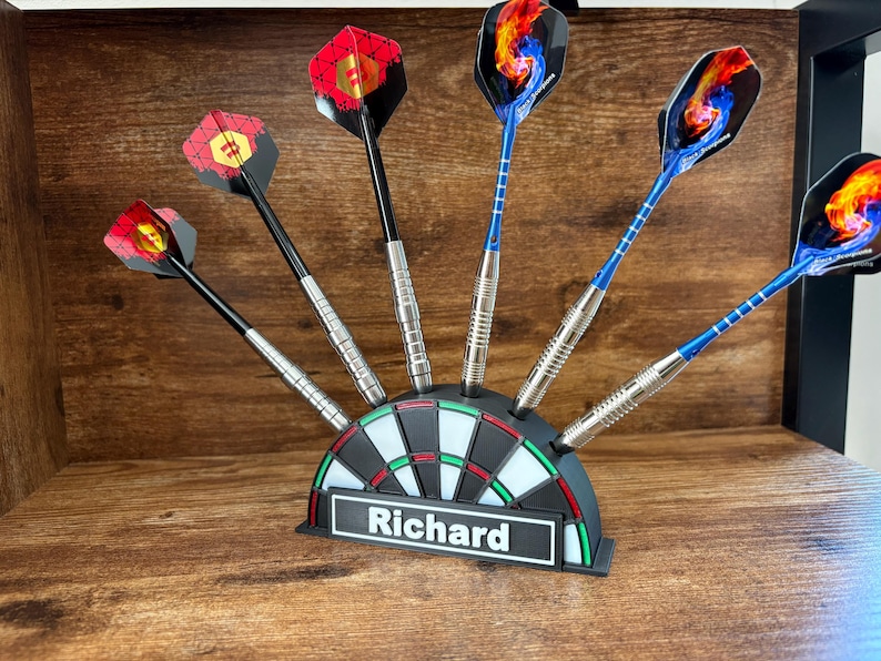 May include: A set of darts with various colorful flights, displayed in a dartboard-shaped holder. The holder is black, white, and green, with the name "Richard" on a white label. The darts have silver barrels and black or blue shafts.