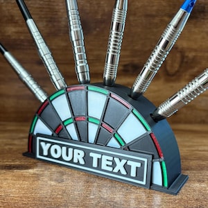 May include: A black and white dartboard-shaped dart holder with silver darts. The holder has a rectangular area with the text "YOUR TEXT" in white. The darts are silver with black and blue accents.