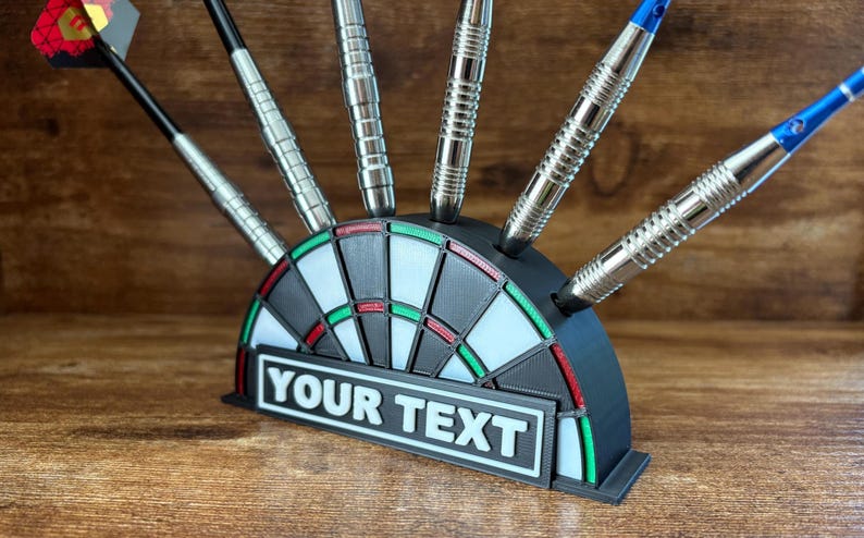 May include: A black and white dartboard-shaped dart holder with silver darts. The holder has a rectangular area with the text "YOUR TEXT" in white. The darts are silver with black and blue accents.