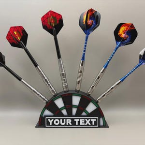 May include: A set of darts with colorful flights, displayed in a dartboard-shaped holder. The darts have silver shafts and black or blue stems. The holder has a black and white target design with the text "YOUR TEXT" on a white rectangle.