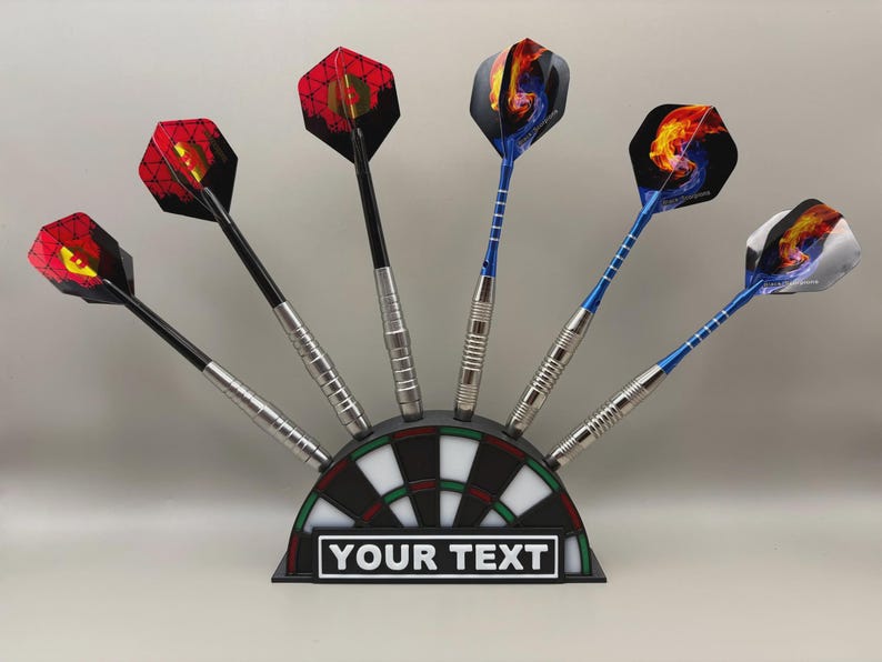 May include: A set of darts with colorful flights, displayed in a dartboard-shaped holder. The darts have silver shafts and black or blue stems. The holder has a black and white target design with the text "YOUR TEXT" on a white rectangle.