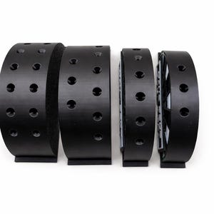 May include: Four black curved objects with evenly spaced holes, possibly parts of a larger mechanism, are arranged in a row against a white background. The objects have a matte finish and a rectangular base.