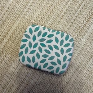 Puede incluir: Una pequeña lata de metal blanca con un patrón de hojas verde azulado. La lata tiene una tapa con bisagras.