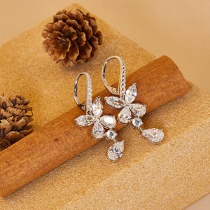 May include: A pair of silver-toned flower-shaped earrings with clear, sparkling gemstones. Each earring features a floral design with multiple petals and a dangling teardrop-shaped gem. The earrings are displayed on a brown surface with pinecones.
