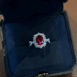 May include: A ring with a large red gemstone surrounded by smaller white gemstones. The ring is set in white metal and has a delicate design.
