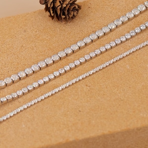 Puede incluir: Primer plano de tres pulseras de tenis plateadas con piedras preciosas claras y brillantes. Las pulseras varían en tamaño, con las piedras preciosas disminuyendo de tamaño. Una piña se encuentra en el fondo. Joyería clásica.
