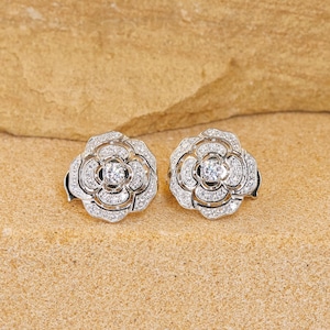 May include: Two silver-coloured stud earrings, each designed as a rose with multiple layers of petals. The centre of each flower features a sparkling, round-cut gemstone, surrounded by smaller, clear stones. The earrings are set against a neutral background.
