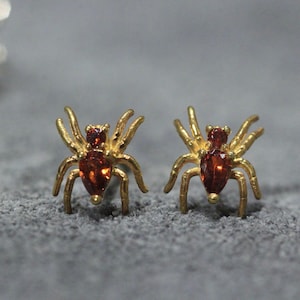 Puede incluir: Un par de pendientes de araña de oro con detalles de piedras preciosas rojas. Las arañas tienen ocho patas y un diseño detallado.