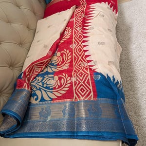 K&ouml;nnte beinhalten: Ein cremefarbener Stoff mit roten und blauen Akzenten. Der Stoff weist komplizierte Muster in Rot und Gold auf, mit einem blauen Rand. Das Design umfasst florale und geometrische Motive, die ein lebendiges und detailliertes Aussehen erzeugen. Der Stoff ist &uuml;ber eine Oberfl&auml;che drapiert.