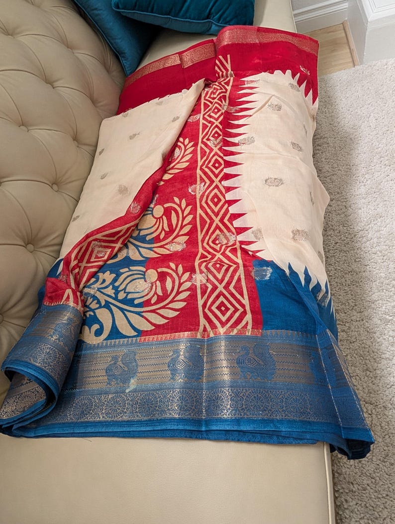 K&ouml;nnte beinhalten: Ein cremefarbener Stoff mit roten und blauen Akzenten. Der Stoff weist komplizierte Muster in Rot und Gold auf, mit einem blauen Rand. Das Design umfasst florale und geometrische Motive, die ein lebendiges und detailliertes Aussehen erzeugen. Der Stoff ist &uuml;ber eine Oberfl&auml;che drapiert.