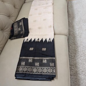 K&ouml;nnte beinhalten: Ein cremefarbener Sari mit goldenen Akzenten und einem schwarzen Rand mit geometrischem Design und Pfauenmotiven. Eine passende schwarze Bluse mit goldenem Rand ist ebenfalls vorhanden. Der Sari liegt gefaltet auf einem beigen Sofa.