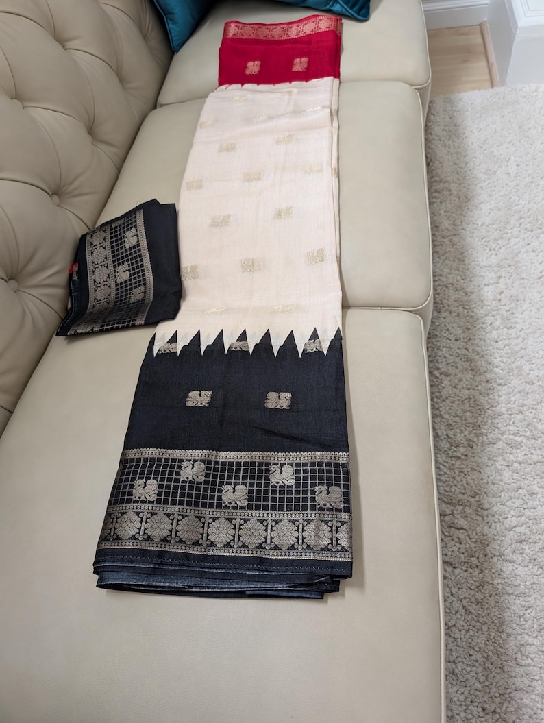 K&ouml;nnte beinhalten: Ein cremefarbener Sari mit goldenen Akzenten und einem schwarzen Rand mit geometrischem Design und Pfauenmotiven. Eine passende schwarze Bluse mit goldenem Rand ist ebenfalls vorhanden. Der Sari liegt gefaltet auf einem beigen Sofa.