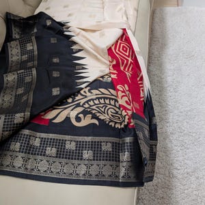 K&ouml;nnte beinhalten: Ein drapiertes Textil mit einem Farbschema aus Schwarz, Creme und Rot. Der Stoff zeigt komplizierte Muster, darunter florale und geometrische Designs, mit silbernen Akzenten. Das Textil ist wahrscheinlich ein Sari oder ein &auml;hnliches Kleidungsst&uuml;ck.