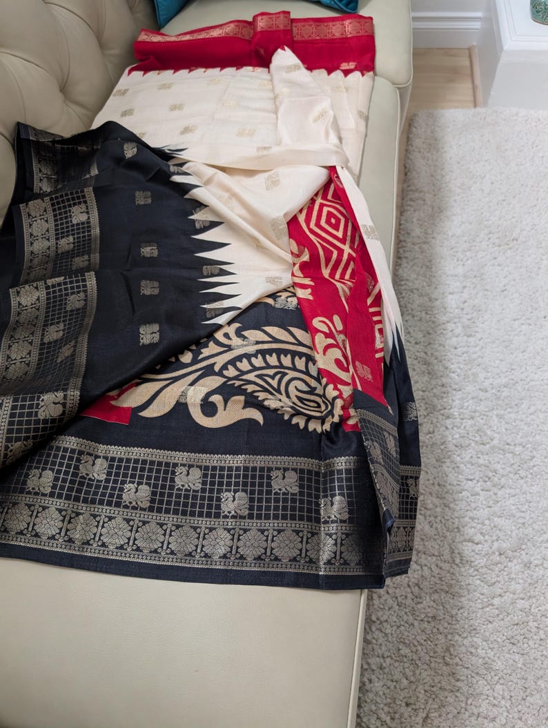 K&ouml;nnte beinhalten: Ein drapiertes Textil mit einem Farbschema aus Schwarz, Creme und Rot. Der Stoff zeigt komplizierte Muster, darunter florale und geometrische Designs, mit silbernen Akzenten. Das Textil ist wahrscheinlich ein Sari oder ein &auml;hnliches Kleidungsst&uuml;ck.