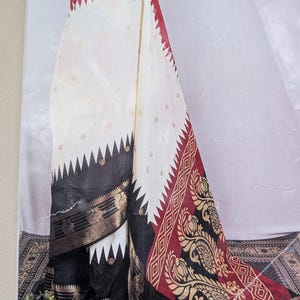 K&ouml;nnte beinhalten: Ein wei&szlig;-roter Sari mit schwarzem Rand. Der Sari hat ein dekoratives Muster mit goldenen Akzenten. Das Design umfasst ein Zickzackmuster an den R&auml;ndern und florale Motive. Der Sari ist &uuml;ber einer Schaufensterpuppe drapiert.