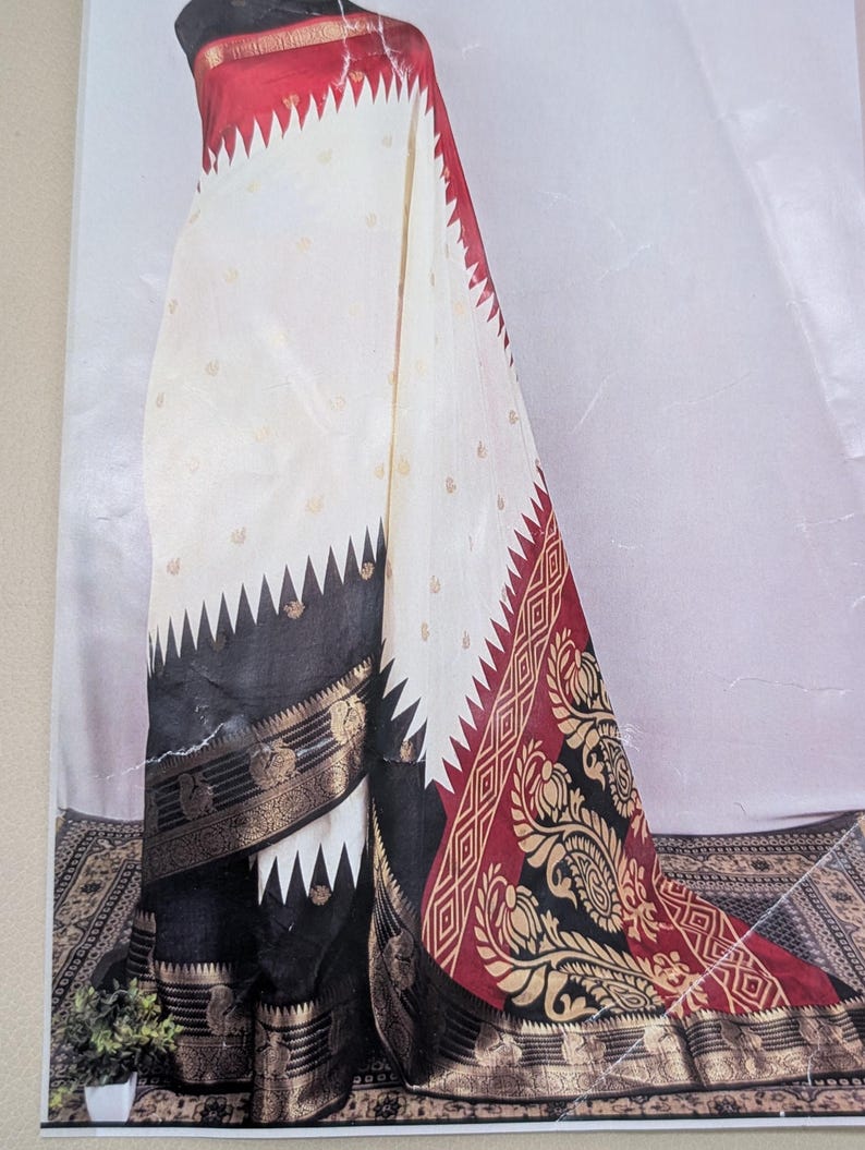 K&ouml;nnte beinhalten: Ein wei&szlig;-roter Sari mit schwarzem Rand. Der Sari hat ein dekoratives Muster mit goldenen Akzenten. Das Design umfasst ein Zickzackmuster an den R&auml;ndern und florale Motive. Der Sari ist &uuml;ber einer Schaufensterpuppe drapiert.
