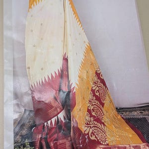 K&ouml;nnte beinhalten: Ein drapiertes Saree mit cremefarbenem K&ouml;rper und kleinen goldenen Akzenten. Der Saum ist tiefrot mit goldenen Verzierungen und einem gelb-goldenen Musterbereich. Das Saree hat ein dekoratives Paisley-Design.