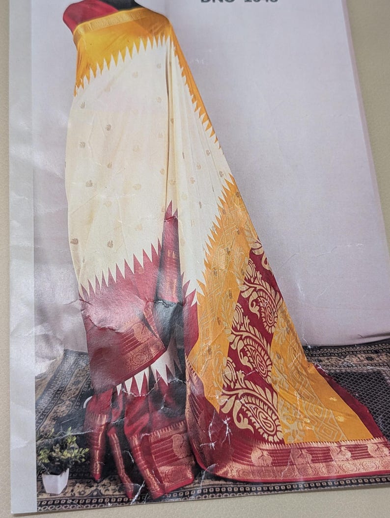 K&ouml;nnte beinhalten: Ein drapiertes Saree mit cremefarbenem K&ouml;rper und kleinen goldenen Akzenten. Der Saum ist tiefrot mit goldenen Verzierungen und einem gelb-goldenen Musterbereich. Das Saree hat ein dekoratives Paisley-Design.