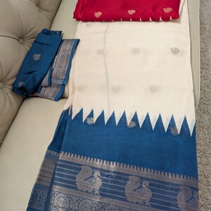 K&ouml;nnte beinhalten: Ein wei&szlig;-blauer Sari mit dekorativem Rand mit Pfauenmotiven. Der Sari hat einen blauen Rand mit Zickzackmuster und ein rotes Stoffst&uuml;ck mit goldenen Akzenten. Der Sari ist auf einer wei&szlig;en Oberfl&auml;che gefaltet.
