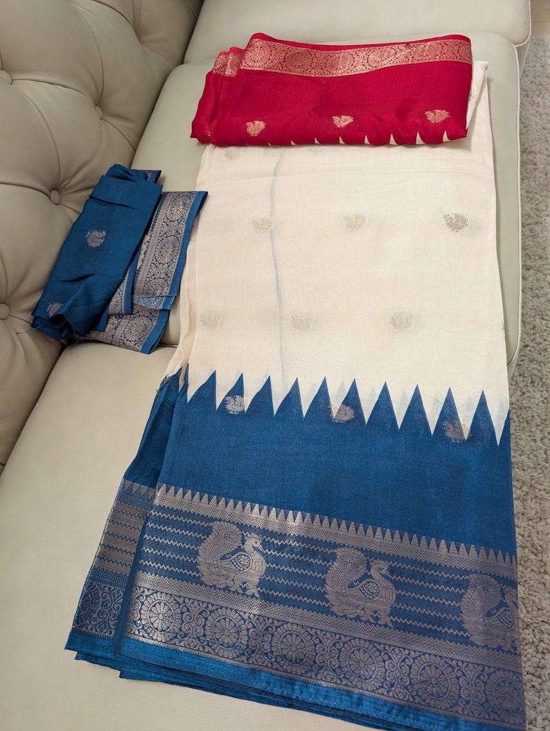 K&ouml;nnte beinhalten: Ein wei&szlig;-blauer Sari mit dekorativem Rand mit Pfauenmotiven. Der Sari hat einen blauen Rand mit Zickzackmuster und ein rotes Stoffst&uuml;ck mit goldenen Akzenten. Der Sari ist auf einer wei&szlig;en Oberfl&auml;che gefaltet.