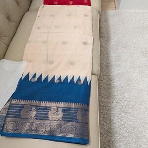 K&ouml;nnte beinhalten: Ein cremefarbenes Textil mit einem blauen Rand mit einem silbernen Pfauenmuster. Das Textil hat einen cremefarbenen K&ouml;rper mit kleinen, sich wiederholenden Mustern. Ein rotes Textil ist im Hintergrund sichtbar.