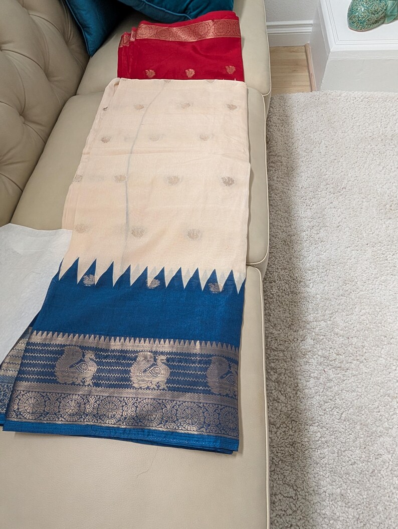 K&ouml;nnte beinhalten: Ein cremefarbenes Textil mit einem blauen Rand mit einem silbernen Pfauenmuster. Das Textil hat einen cremefarbenen K&ouml;rper mit kleinen, sich wiederholenden Mustern. Ein rotes Textil ist im Hintergrund sichtbar.