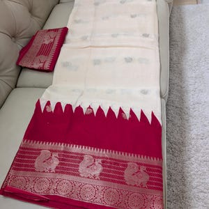 K&ouml;nnte beinhalten: Ein cremefarbener Sari mit einem leuchtend roten Rand. Der Rand weist aufwendige silberne Pfauen- und Blumendesigns auf. Der Sari ist &uuml;ber einem wei&szlig;en Sofa drapiert und zeigt seine elegante Textur und sein Design.