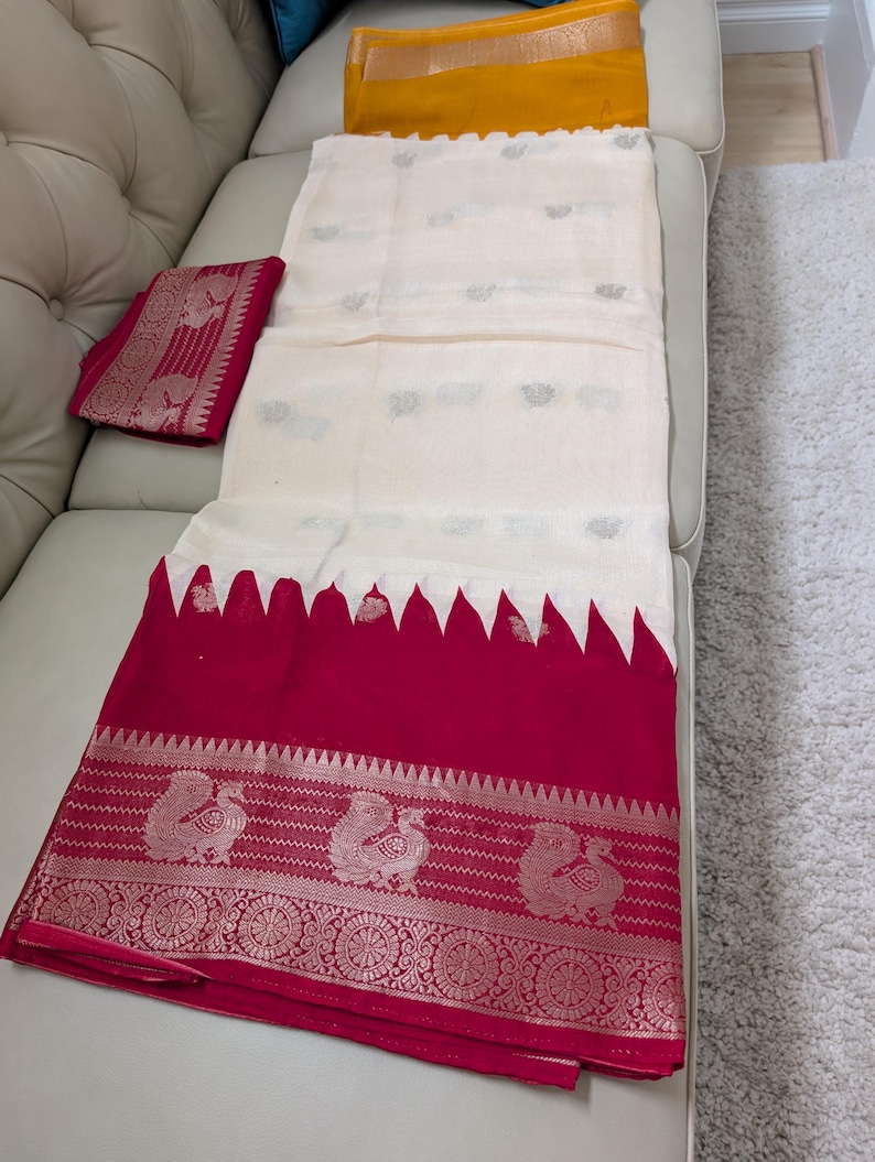 K&ouml;nnte beinhalten: Ein cremefarbener Sari mit einem leuchtend roten Rand. Der Rand weist aufwendige silberne Pfauen- und Blumendesigns auf. Der Sari ist &uuml;ber einem wei&szlig;en Sofa drapiert und zeigt seine elegante Textur und sein Design.