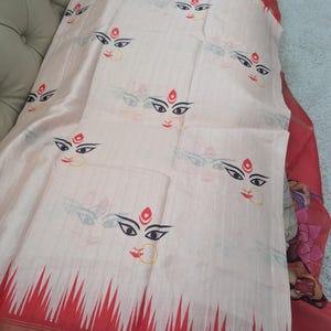 May include: A white silk saree with a red border and a repeating pattern of a woman's face. The woman's face is painted in black, red, and gold.