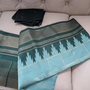 May include: A teal and green textile with intricate patterns and a matching dark green fabric square. The textile features geometric designs and a light blue border. The fabric is draped over a white surface.