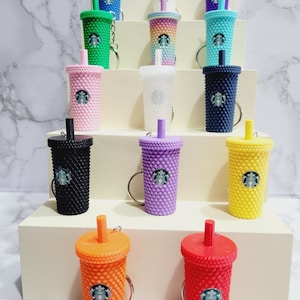 Könnte beinhalten: Eine Sammlung von 14 bunten Starbucks-Becher-Schlüsselanhängern. Die Anhänger sind aus Kunststoff und haben ein strukturiertes Design. Die Anhänger sind pyramidenförmig auf einer weißen Oberfläche angeordnet.