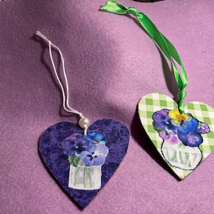 Puede incluir: Dos adornos de madera en forma de corazón con diseños florales. Un adorno es morado con un ramo de pensamientos morados y blancos en un jarrón transparente. El otro adorno es de cuadros verdes y blancos con un ramo de pensamientos morados y amarillos en un jarrón blanco.