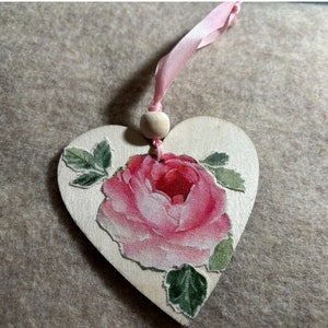 May include: A wooden heart-shaped ornament with a pink rose and green leaves printed on it. The ornament has a pink ribbon attached to it.