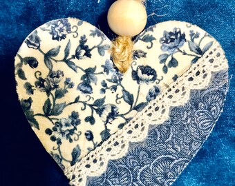 Op de afbeelding: Een hartvormige stoffen ornament met een blauw en wit bloemenpatroon. De ornament is versierd met witte kant en heeft een houten kraal aan de bovenkant.