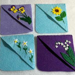 Puede incluir: Cuatro marcapáginas de fieltro de esquina con flores bordadas. Los marcapáginas son morados, azules y verdes. Las flores son pensamientos amarillos, un girasol amarillo, margaritas blancas y lirios del valle blancos.