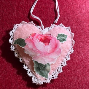 May include: A pink felt heart-shaped ornament with a white lace trim and a pink rose with green leaves printed on the front.
