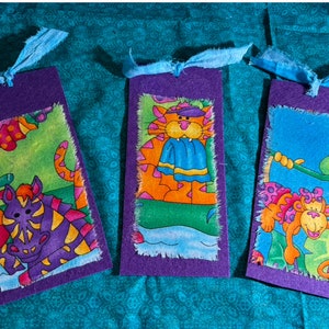 May include: Three purple fabric bookmarks with colorful animal illustrations. The first bookmark features a purple and yellow zebra, the second bookmark features an orange cat wearing a blue scarf, and the third bookmark features a pink and orange monkey hanging from a branch.