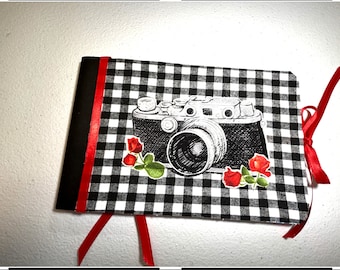 Könnte beinhalten: Ein Fotoalbum aus schwarzem und weißem kariertem Stoff mit einem roten Bandverschluss. Das Album zeigt eine Illustration einer Vintage-Kamera mit roten Rosen.