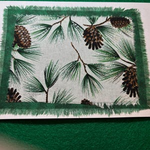 May include: A white card with a green border and a printed image of pine branches and pine cones. The pine cones are brown and have a glittery finish.