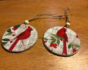 May include: Two round, fabric ornaments with a red cardinal bird on each. The ornaments are decorated with a white background and a musical note pattern. Each ornament has a small wooden bead and twine for hanging.