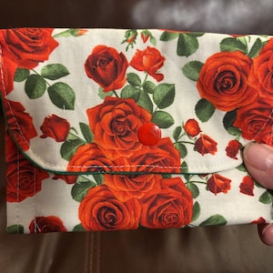 May include: A rectangular fabric wallet featuring a vibrant floral pattern of red roses and green leaves on an off-white background. The wallet has a fold-over flap secured with a single red plastic snap button and finished with green trim.