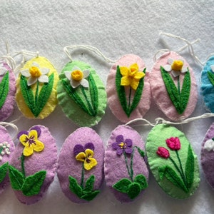 Puede incluir: Doce adornos de huevos de Pascua de fieltro, cada uno decorado con una flor de primavera diferente. Las flores incluyen narcisos, pensamientos, tulipanes y lirios del valle. Los huevos son de varios colores pastel, incluyendo morado, rosa, amarillo, verde y azul.