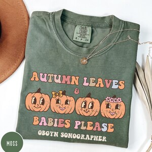 May include: A moss green t-shirt with the text "AUTUMN LEAVES & BABIES PLEASE OBGYN SONOGRAPHER" and four cartoon pumpkins. The shirt has a crew neck and short sleeves. A gold necklace is draped across the shirt.