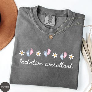 May include: A gray Comfort Colors t-shirt with the words "lactation consultant" in white script. The shirt features a design of white daisies and baby footprints in pink and blue. A silver necklace with a pendant is draped across the shirt.